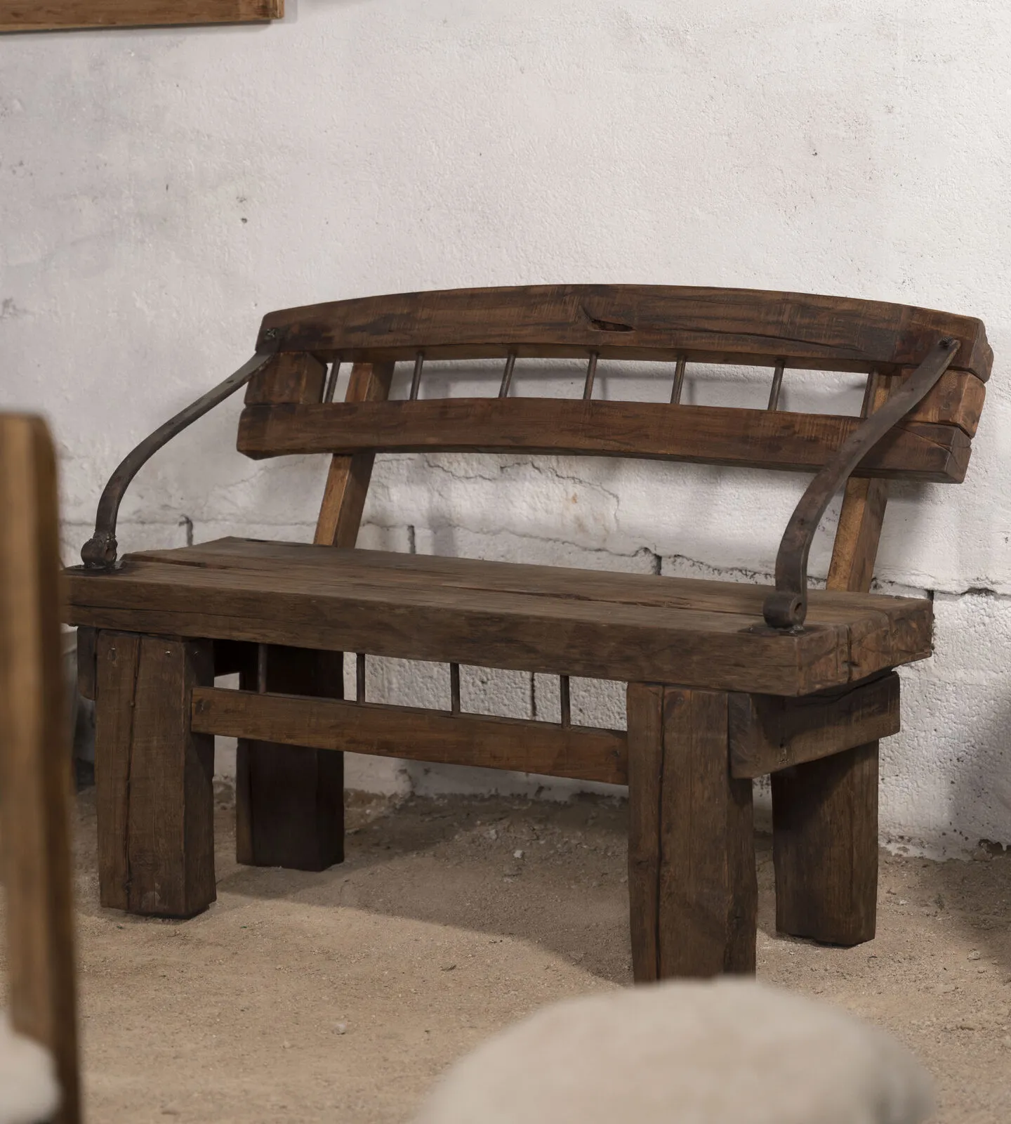 Banco de madera oscura con respaldo de barrotes y brazos de hierro curvado, junto a una lechera